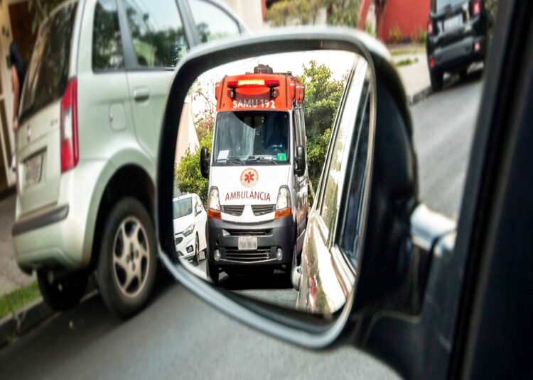 Ambulância branca do SAMU 192 com sirene ligada refletida no espelho retrovisor de um veículo