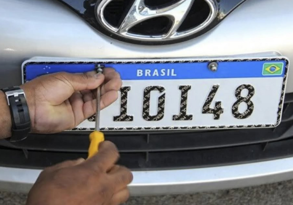Mãos masculinas instalam placa Mercosul com código IDI48 na dianteira de veículo Hyundai prata usando chave de fenda