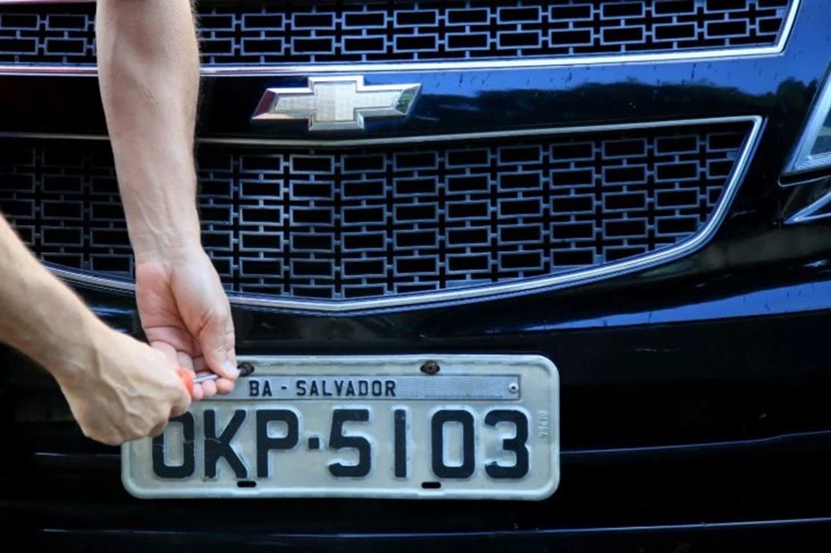 Mão segurando placa cinza de carro OKP-5103 com inscrição BA-Salvador na frente de Chevrolet preto