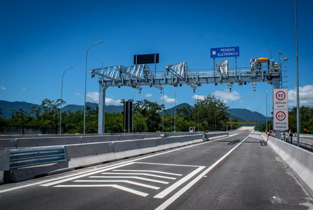 Pórtico metálico de pedágio eletrônico sobre rodovia com placa azul "Pedágio Eletrônico" e limites de velocidade