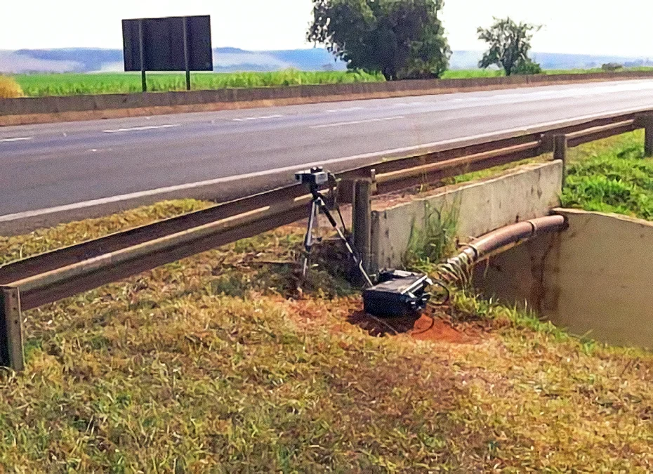 Radar portátil em tripé atrás de guard rail metálico em rodovia com vegetação verde ao fundo