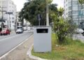 Caixa metálica cinza em canteiro central de avenida urbana, usada como radar de velocidade camuflado