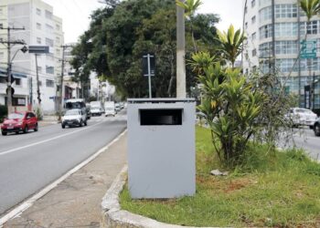 Caixa metálica cinza em canteiro central de avenida urbana, usada como radar de velocidade camuflado