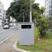 Caixa metálica cinza em canteiro central de avenida urbana, usada como radar de velocidade camuflado
