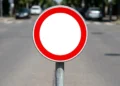 Placa circular com borda vermelha e centro branco sem símbolo, fixada em poste metálico em via urbana com carros ao fundo