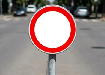 Placa circular com borda vermelha e centro branco sem símbolo, fixada em poste metálico em via urbana com carros ao fundo