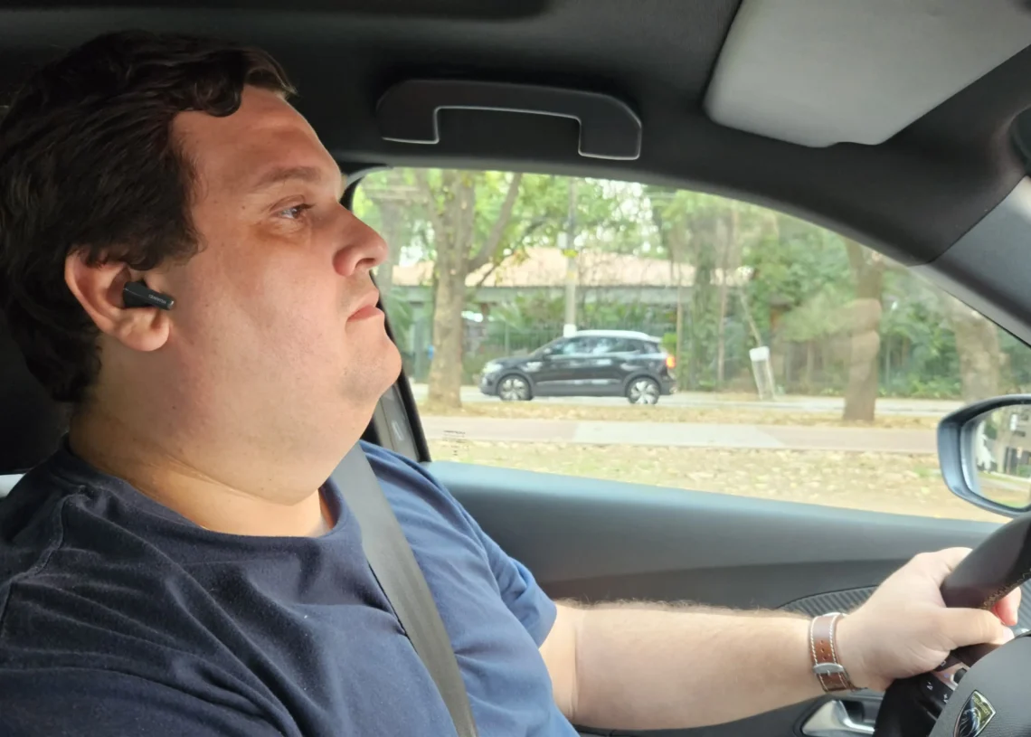 Homem de camiseta azul com fone bluetooth preto no ouvido esquerdo dirigindo veículo com cinto afivelado