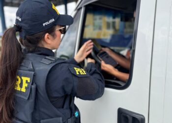 Policial rodoviário federal com uniforme azul e boné PRF aborda condutor de caminhão branco, segurando tablet