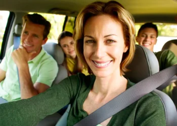 Mulher sorridente dirige carro com família ao fundo, usando cinto de segurança em dia ensolarado de viagem