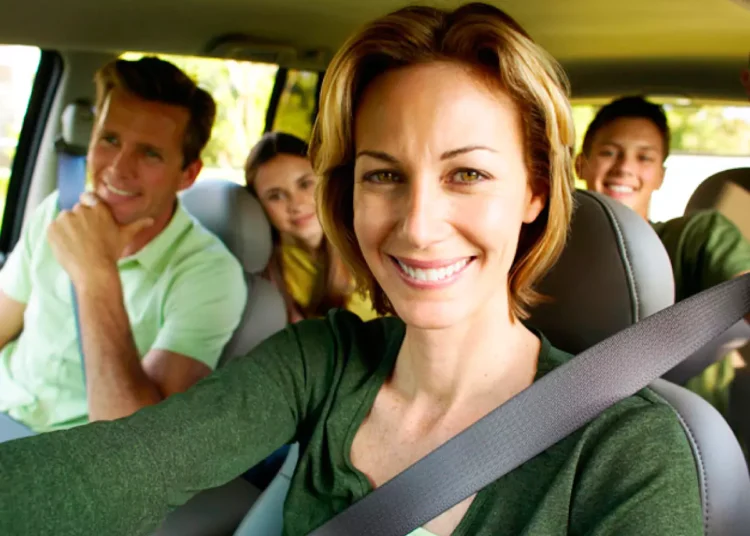 Mulher sorridente dirige carro com família ao fundo, usando cinto de segurança em dia ensolarado de viagem