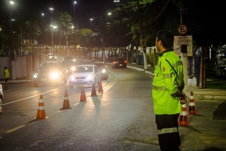 Agente de trânsito com colete refletivo verde fiscaliza via noturna com cones laranjas e veículos com faróis acesos.