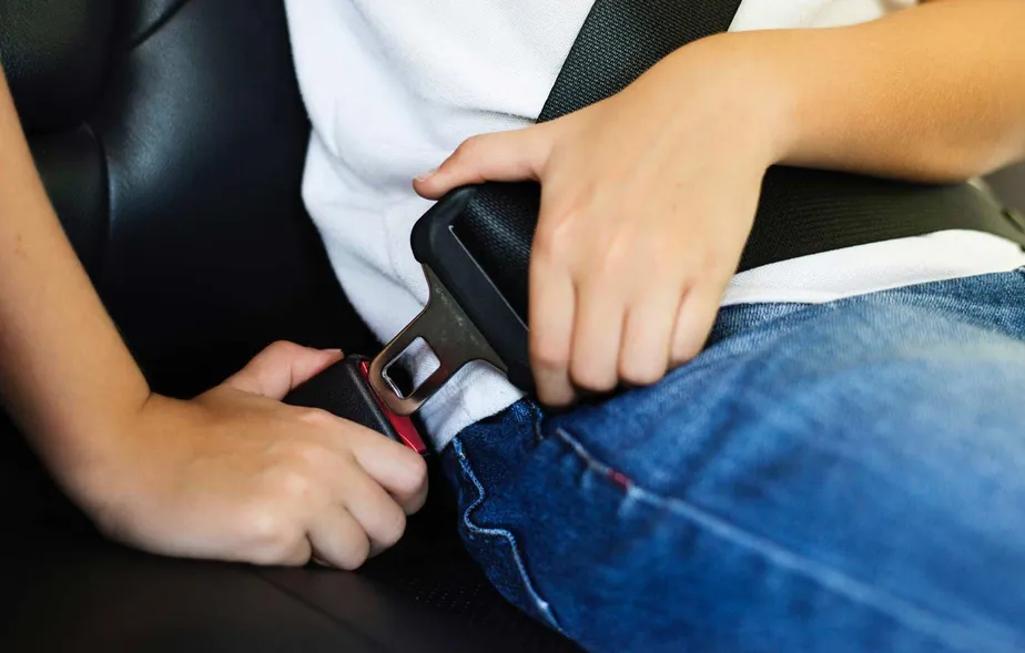 Mãos afivelando cinto de segurança preto em banco de carro, pessoa usando camiseta branca e calça jeans