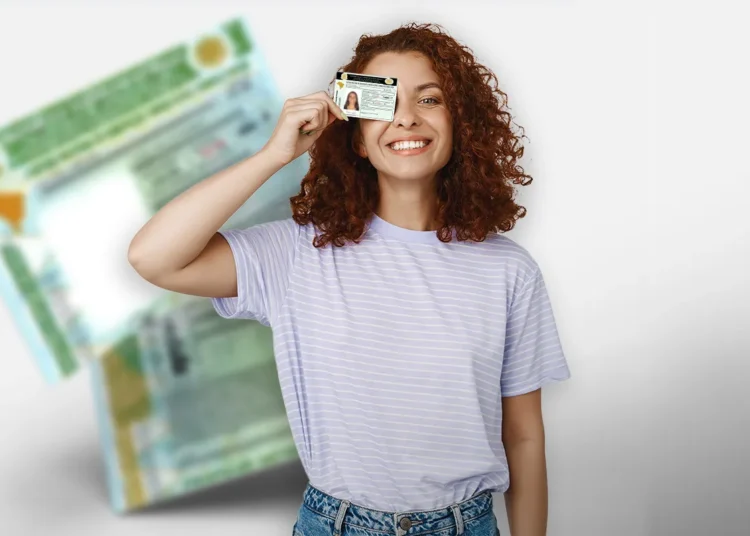 Mulher de cabelos ruivos cacheados e camiseta lilás listrada segura CNH sobre o olho esquerdo, sorrindo.