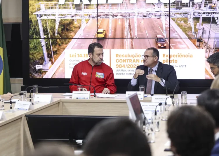 Guilherme Boulos de jaqueta vermelha e George Santoro de terno escuro em coletiva sobre pedágios eletrônicos