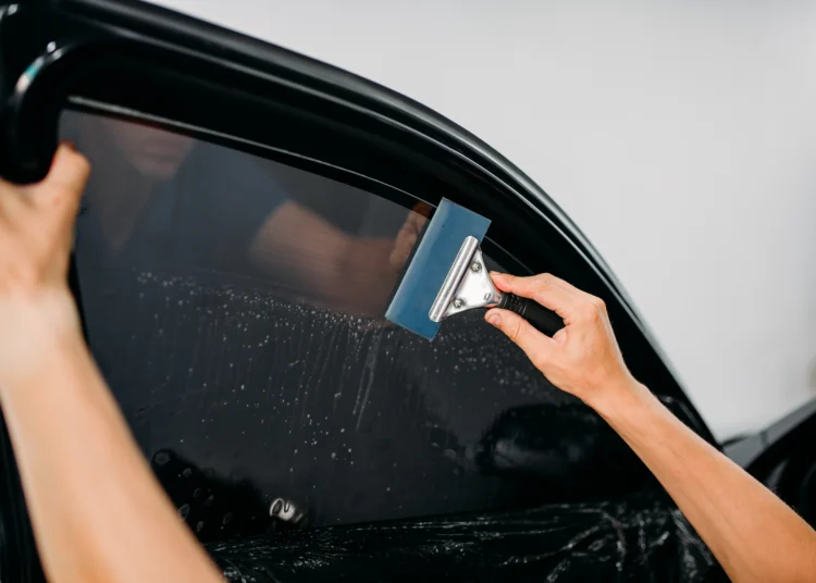 Profissional aplica película escura no vidro lateral de um carro preto, utilizando um rodo azul para alisar o filme e remover bolhas de ar e água.