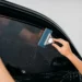 Profissional aplica película escura no vidro lateral de um carro preto, utilizando um rodo azul para alisar o filme e remover bolhas de ar e água.