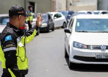 Agente de trânsito com uniforme amarelo e boné ergue a mão para orientar veículos em rua de Salvador.