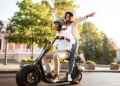 Casal sorridente pilota scooter elétrica preta com design moderno, ambos usando capacete em área urbana arborizada.