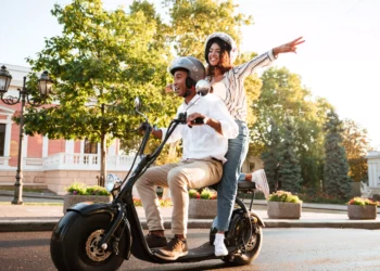 Casal sorridente pilota scooter elétrica preta com design moderno, ambos usando capacete em área urbana arborizada.