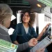 Mulher de cabelos cacheados ao volante sorri enquanto instrutor de cabelo grisalho com prancheta avalia seu desempenho no exame.