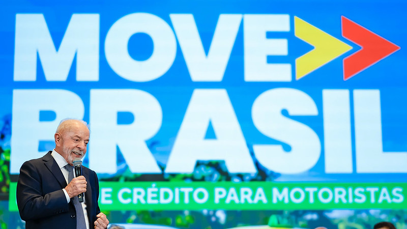 Lula discursa segurando microfone diante de painel azul do Move Brasil com setas coloridas e texto sobre crédito para motoristas