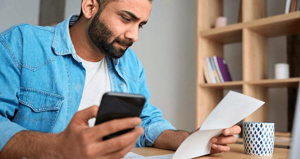 Homem de barba com camisa jeans azul segura celular e papel branco em escritório com estante