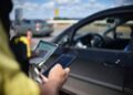 Agente de trânsito com celular e CRLV verifica documentação de veículo escuro em blitz sob céu azul