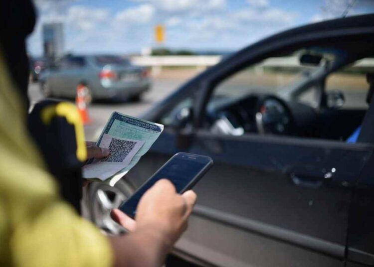 Agente de trânsito com celular e CRLV verifica documentação de veículo escuro em blitz sob céu azul