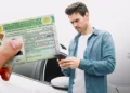 Homem de camisa jeans olha celular ao lado de carro enquanto mão exibe CNH brasileira categoria D
