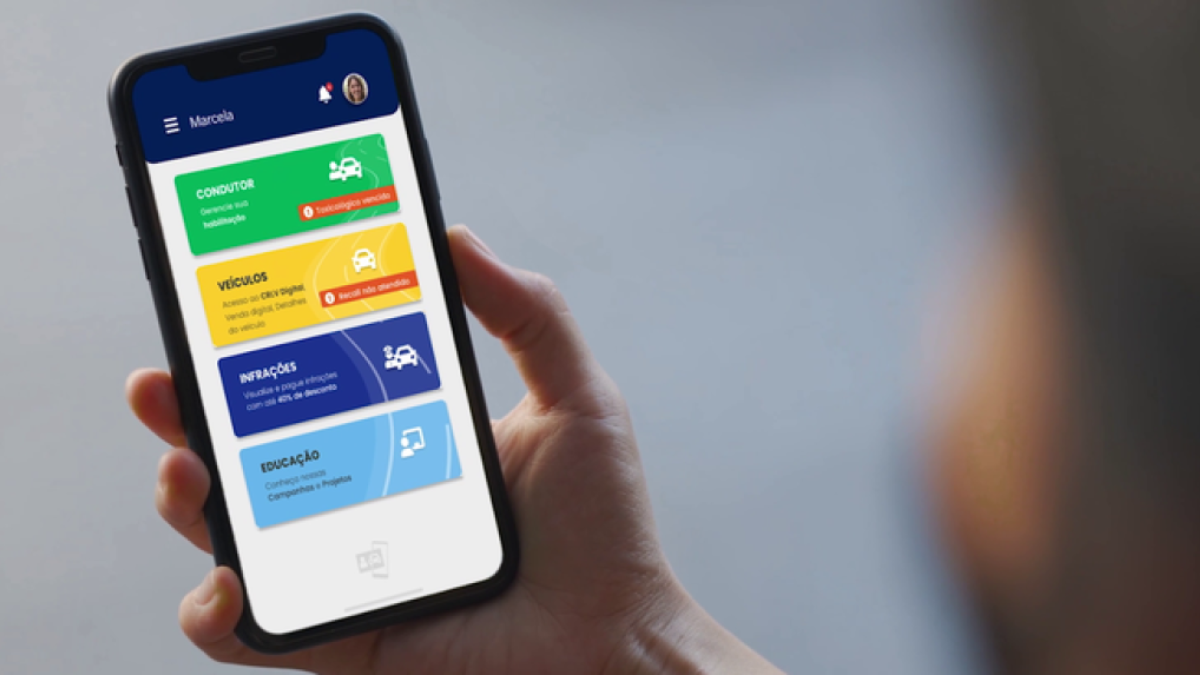 Mão segurando smartphone com aplicativo CNH do Brasil exibindo menu colorido com seções Condutor, Veículos, Infrações e Educação