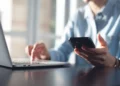 Pessoa de camisa azul segura smartphone preto enquanto digita em notebook prata sobre mesa escura