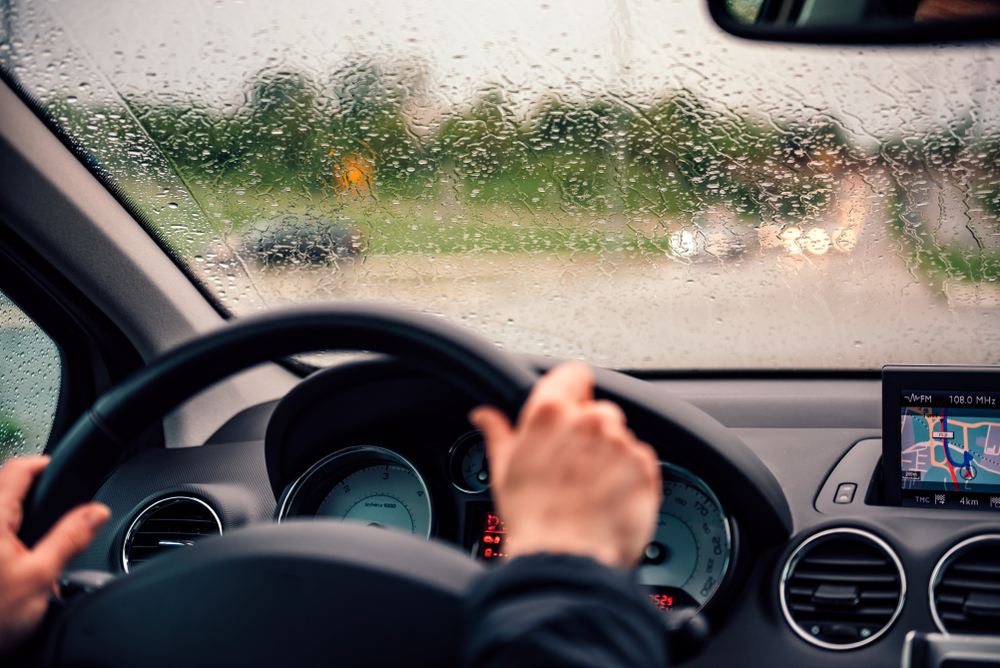 Mãos no volante, dirigindo carro sob chuva forte, painel com velocímetro e tela de GPS exibindo mapa de rota