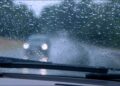 Visão do interior de um carro com gotas de chuva no para-brisa e faróis desfocados de veículo à frente
