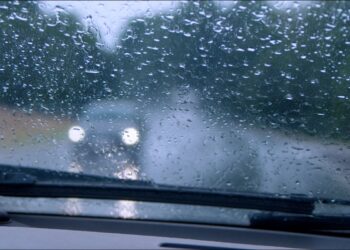 Visão do interior de um carro com gotas de chuva no para-brisa e faróis desfocados de veículo à frente