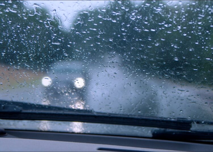Visão do interior de um carro com gotas de chuva no para-brisa e faróis desfocados de veículo à frente