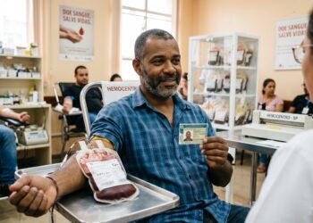 Homem negro de barba grisalha, camisa xadrez azul, doa sangue tipo O+ enquanto mostra CNH para profissional de saúde