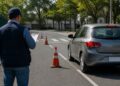 Examinador com boné e prancheta observa carro prata realizando manobra entre cones laranjas em pista de teste