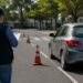 Examinador com boné e prancheta observa carro prata realizando manobra entre cones laranjas em pista de teste