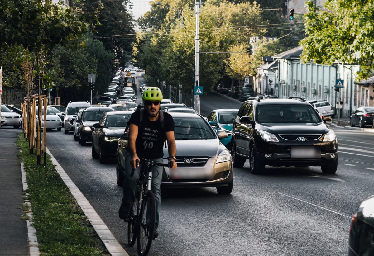 Ciclista com capacete verde e óculos escuros pedala entre fila de carros em via urbana arborizada