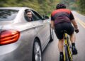 Motorista gritando pela janela de sedã prata ao ultrapassar ciclista em bicicleta amarela em estrada arborizada