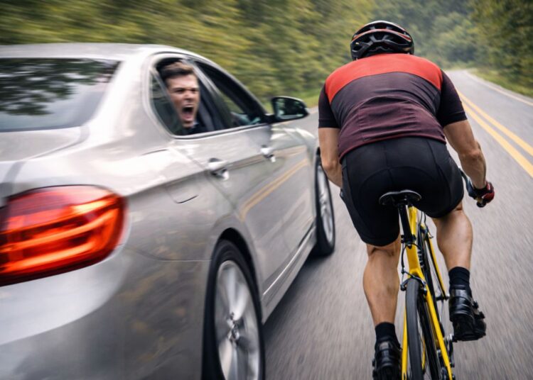 Motorista gritando pela janela de sedã prata ao ultrapassar ciclista em bicicleta amarela em estrada arborizada