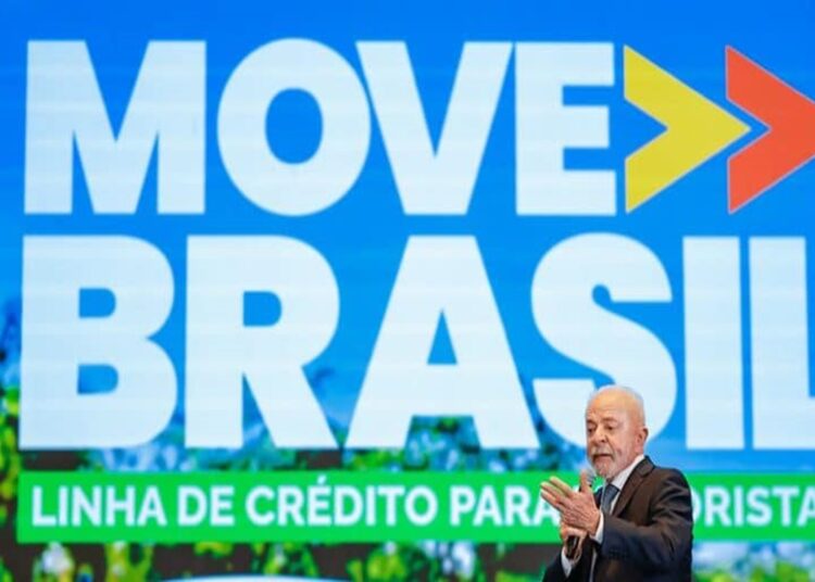 Presidente Lula de terno escuro aplaude em evento do Move Brasil com painel azul ao fundo exibindo logo e texto do programa.