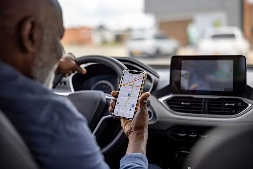 Homem negro de camisa azul dentro de carro segura celular com aplicativo de navegação, painel e multimídia ao fundo
