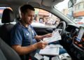 Motorista de aplicativo com camisa polo azul analisa documentos do carnê-leão IRPF dentro do carro com celular no suporte