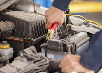 Mão de mecânico com uniforme azul conectando cabo vermelho com garra dourada no terminal positivo de bateria automotiva