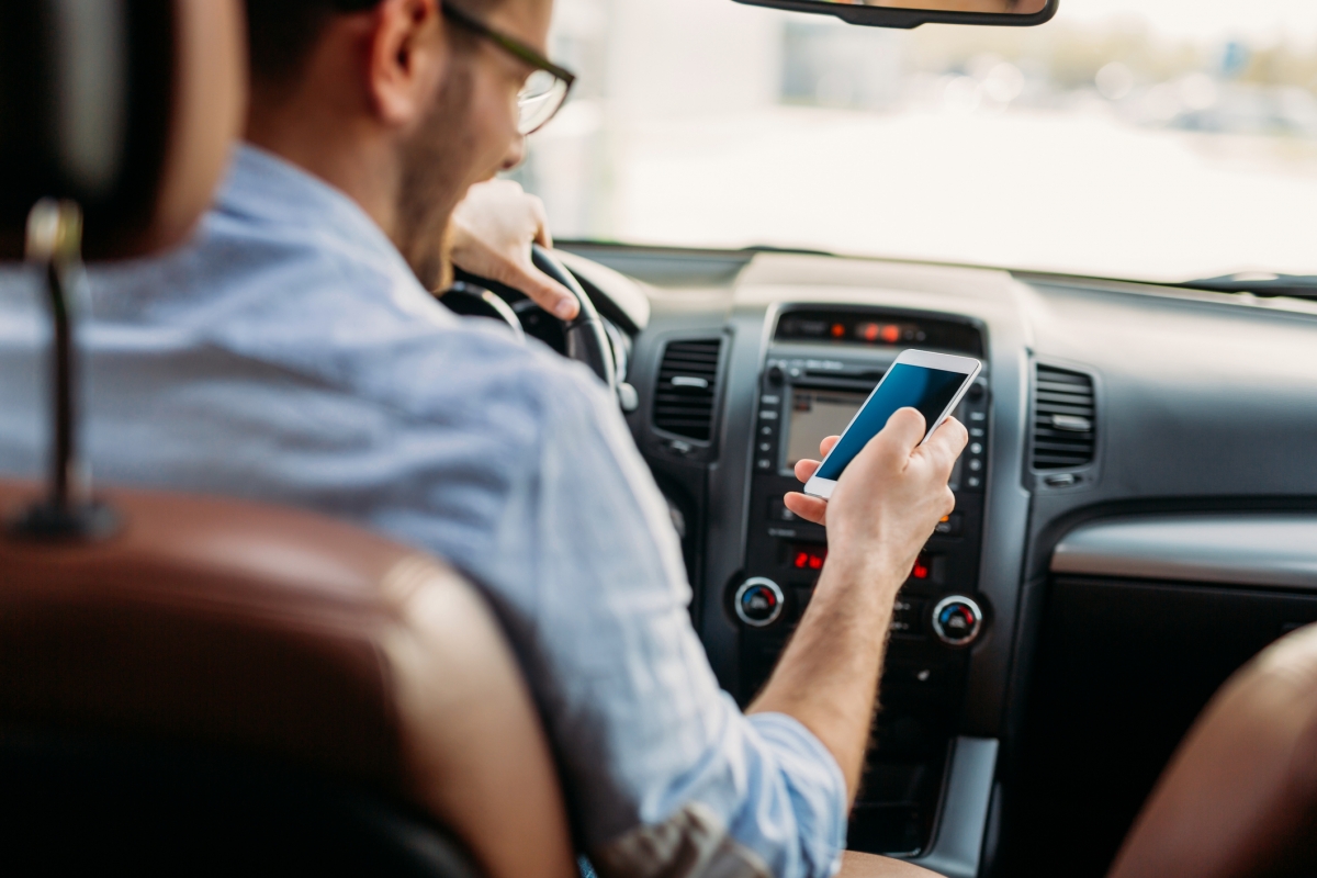 Homem de óculos e camisa azul clara manuseia smartphone branco enquanto dirige veículo com interior marrom