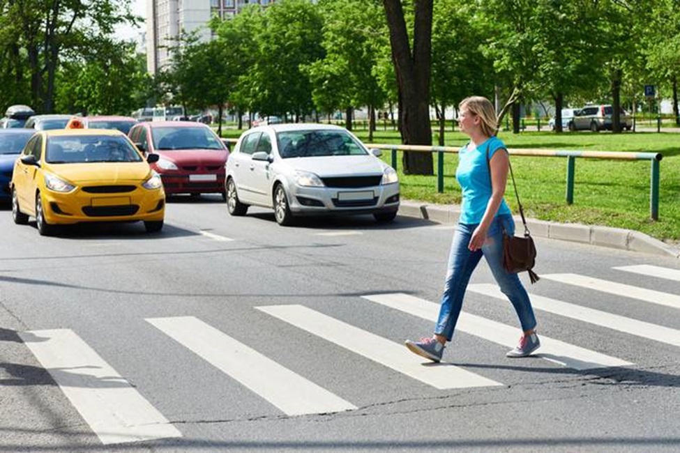 Mulher de blusa azul atravessando faixa de pedestres, com táxi amarelo e carros parados ao fundo