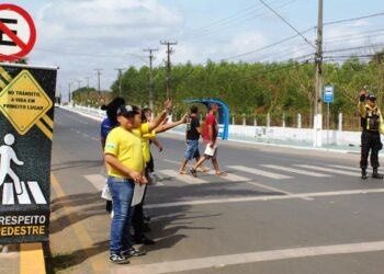 Agentes de trânsito orientam pedestres e motoristas em faixa de segurança com placa "Respeito ao Pedestre"