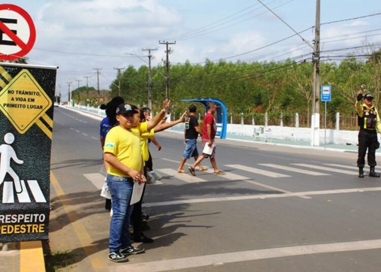 Agentes de trânsito orientam pedestres e motoristas em faixa de segurança com placa "Respeito ao Pedestre"
