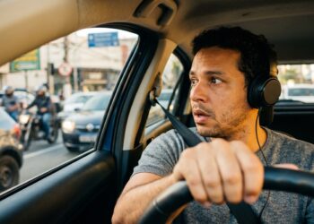 Homem de cabelos cacheados usando fone de ouvido enquanto dirige em trânsito intenso com motos e carros